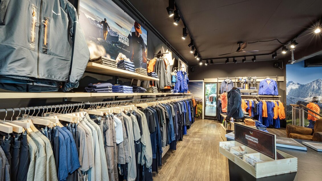 Ein moderner Bekleidungsladen mit einer Vielzahl von sportlicher Kleidung an Regalen. Die Wände sind mit Bildern von Outdoor-Aktivitäten dekoriert. | © Mountain Shop Schladming