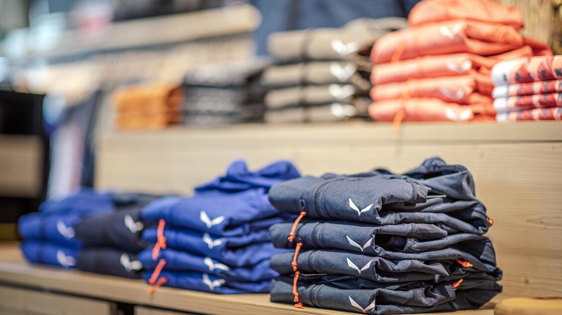 Stapelhaufen von T-Shirts in verschiedenen Farben sind auf einem Holzregal angeordnet. Im Hintergrund sind weitere Kleidungsstücke sichtbar. | © Mountain Shop Schladming