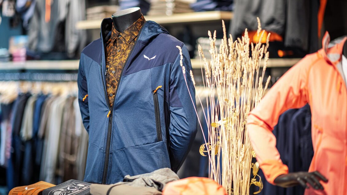 Ein modisches Outfit aus einer blauen Jacke und einem gemusterten Hemd ist auf einem Mannequin ausgestellt. Im Hintergrund sind weitere Kleidungsteile und Accessoires zu sehen. | © Mountain Shop Schladming