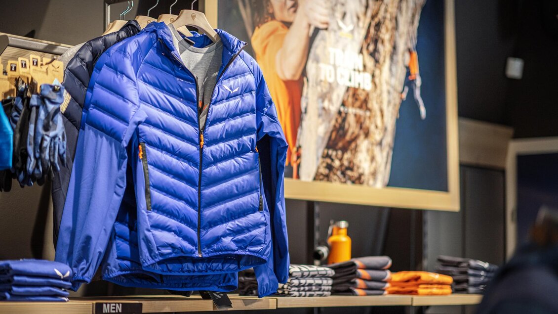 Eine blaue Jacke hängt in einem Sportgeschäft, umgeben von anderen Outdoor-Bekleidungsstücken. Im Hintergrund ist ein Plakat mit einem Kletterer zu sehen. | © Mountain Shop Schladming
