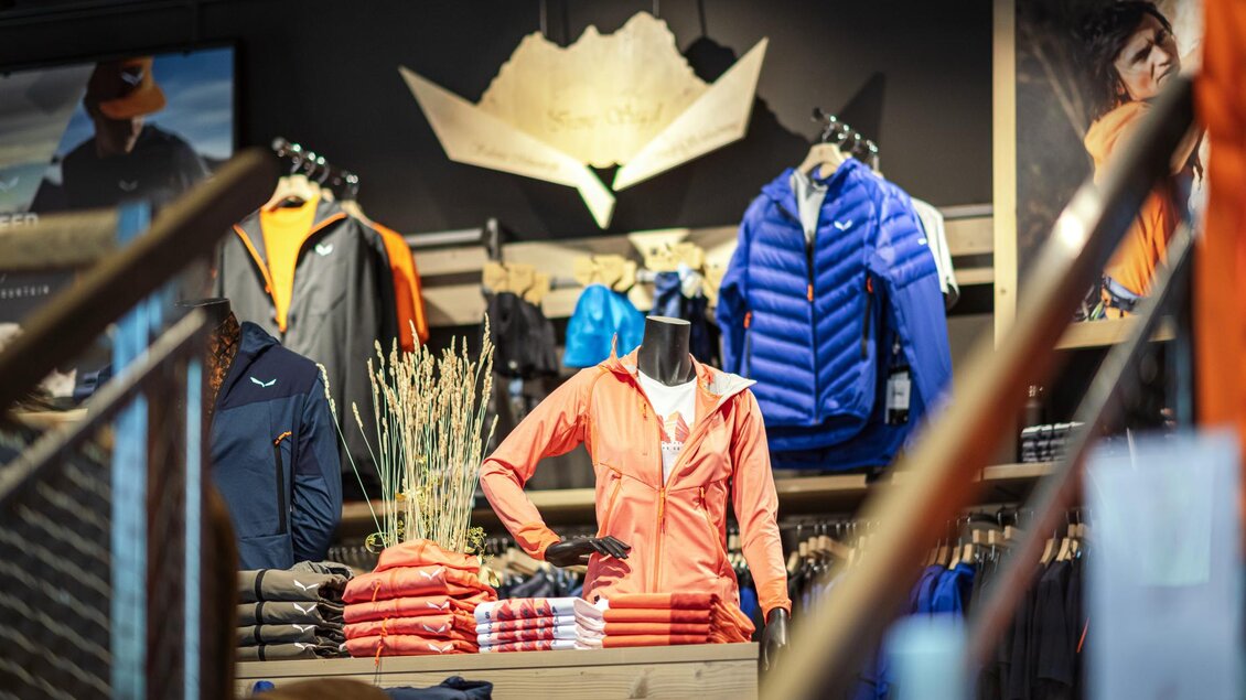 Ein moderner Sportbekleidungsladen mit einer Vielzahl von Jacken und T-Shirts. Im Vordergrund steht ein orangefarbener Anorak an einer Kleiderpuppe. | © Mountain Shop Schladming