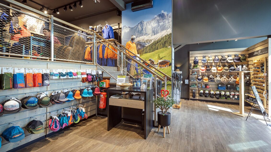 Ein gut sortierter Outdoor-Shop mit verschiedenen Bekleidungs- und Zubehörangeboten. Im Hintergrund sind Bergbilder und eine Treppe zu sehen, die nach oben führt. | © Mountain Shop Schladming