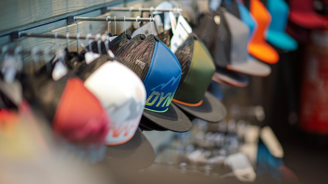 Eine Reihe von verschiedenen Caps hängt an einem Regal. Die Hüte haben unterschiedliche Farben und Designs. | © Mountain Shop Schladming