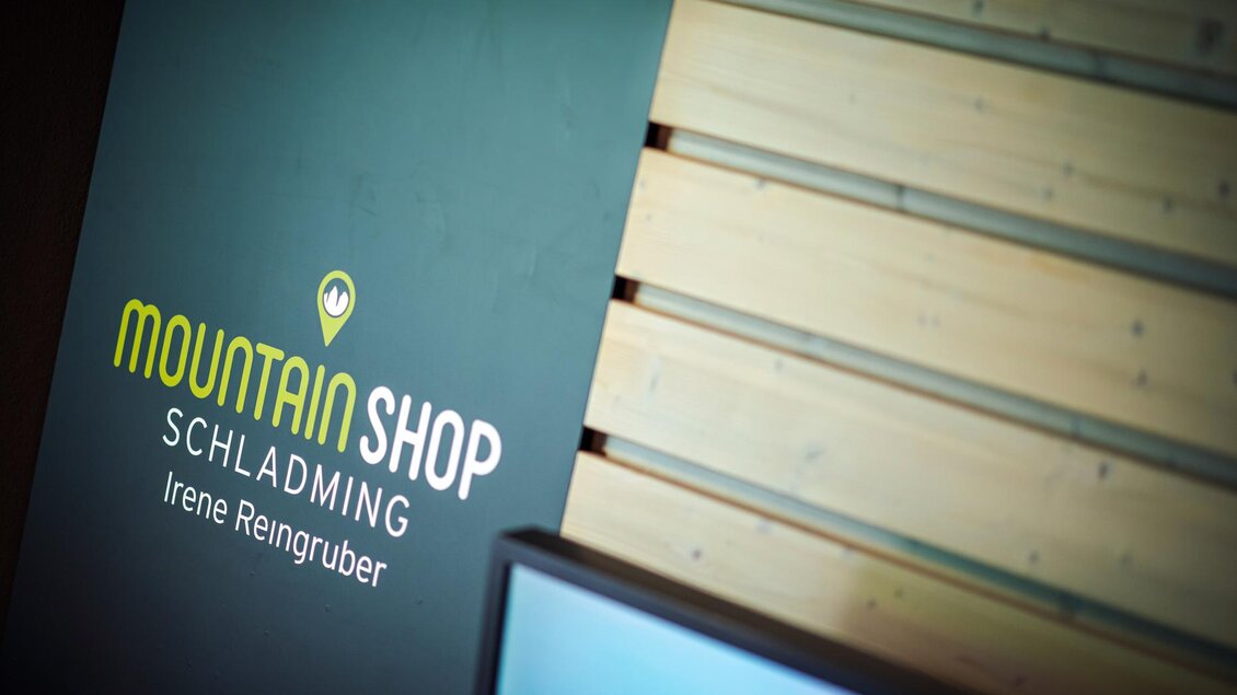 Ein Schild mit der Aufschrift „Mountain Shop Schladming“ ist vor einer holzverkleideten Wand zu sehen. Der Text ist gut lesbar und betont die Verbindung zur Region. | © Mountain Shop Schladming