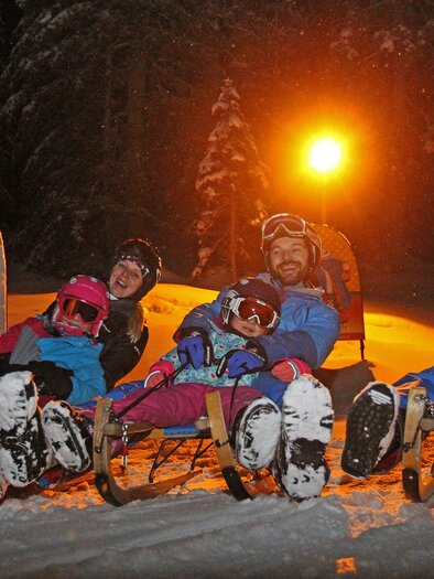 A group of four people is sitting on sleds in the snow. It is dark, and a light glow illuminates the scene. | © Hans-Peter Steiner