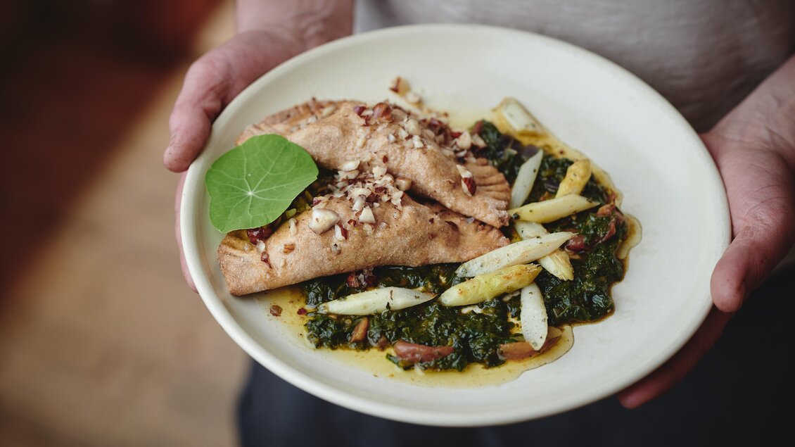 Ein Teller mit gefüllten Teigstücken, garniert mit Nüssen und frischem Blatt. Daneben liegt eine Portion Blattgemüse und Stücke von zarten Spargel. | © Liebstöckel
