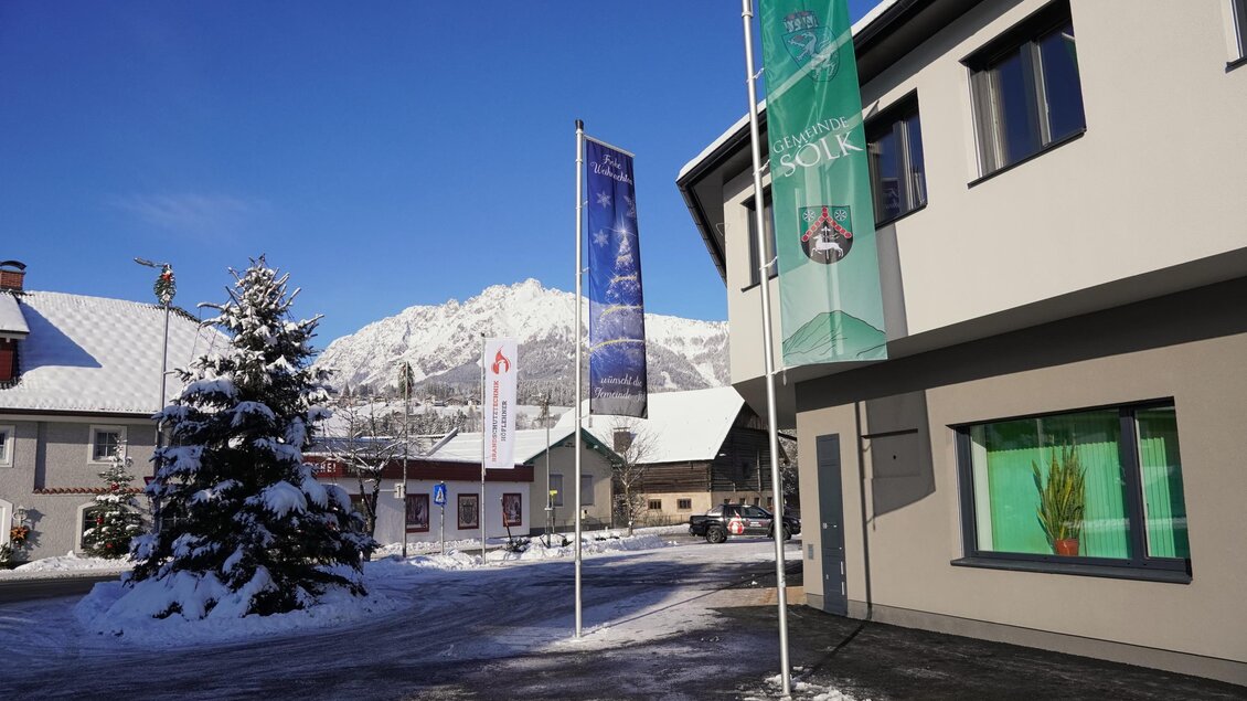 Ein schneebedeckter Ort mit modernen Gebäuden und Fahnen im Vordergrund. Im Hintergrund sind Berge unter einem klaren blauen Himmel zu sehen. | © Gemeinde Sölk
