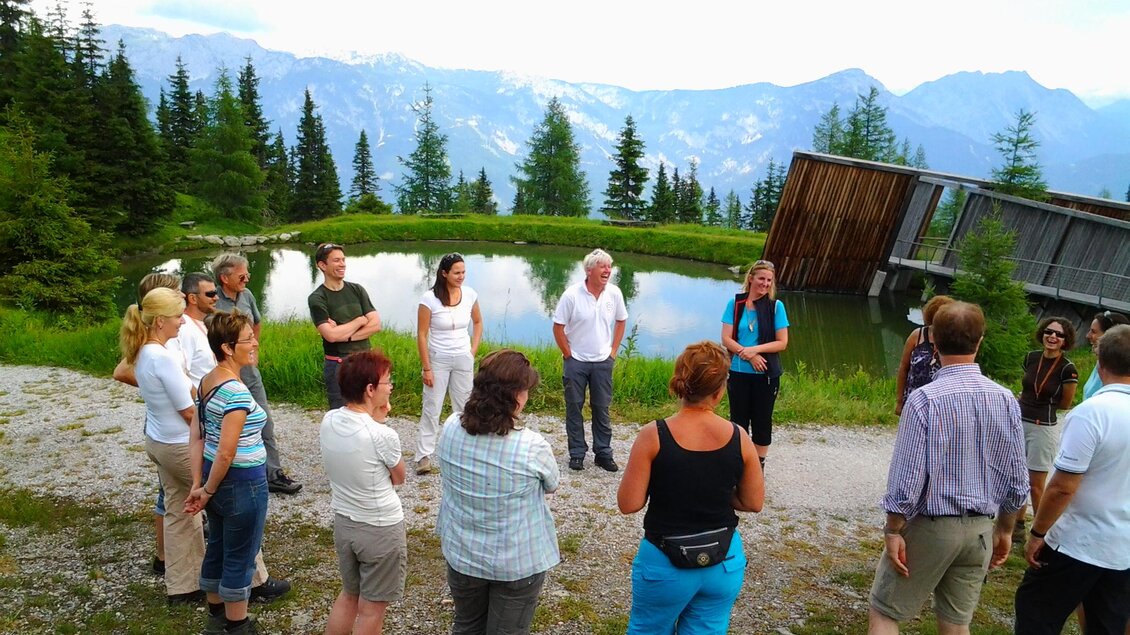 Eine Gruppe von Menschen versammelt sich in der Natur um einen Teich. Im Hintergrund sind Berge und Bäume sichtbar. | © Thomas Humer