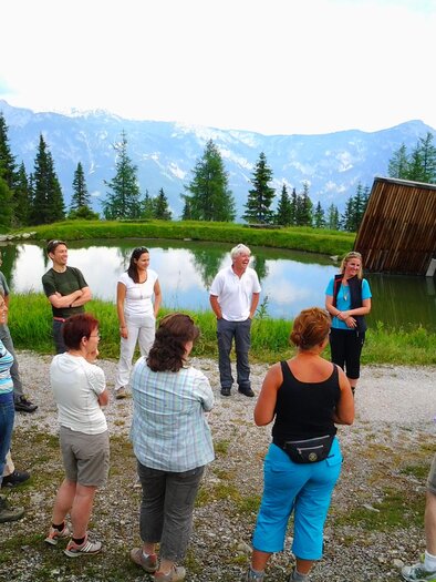 A group of people gathers in nature around a pond. In the background, mountains and trees are visible. | © Thomas Humer