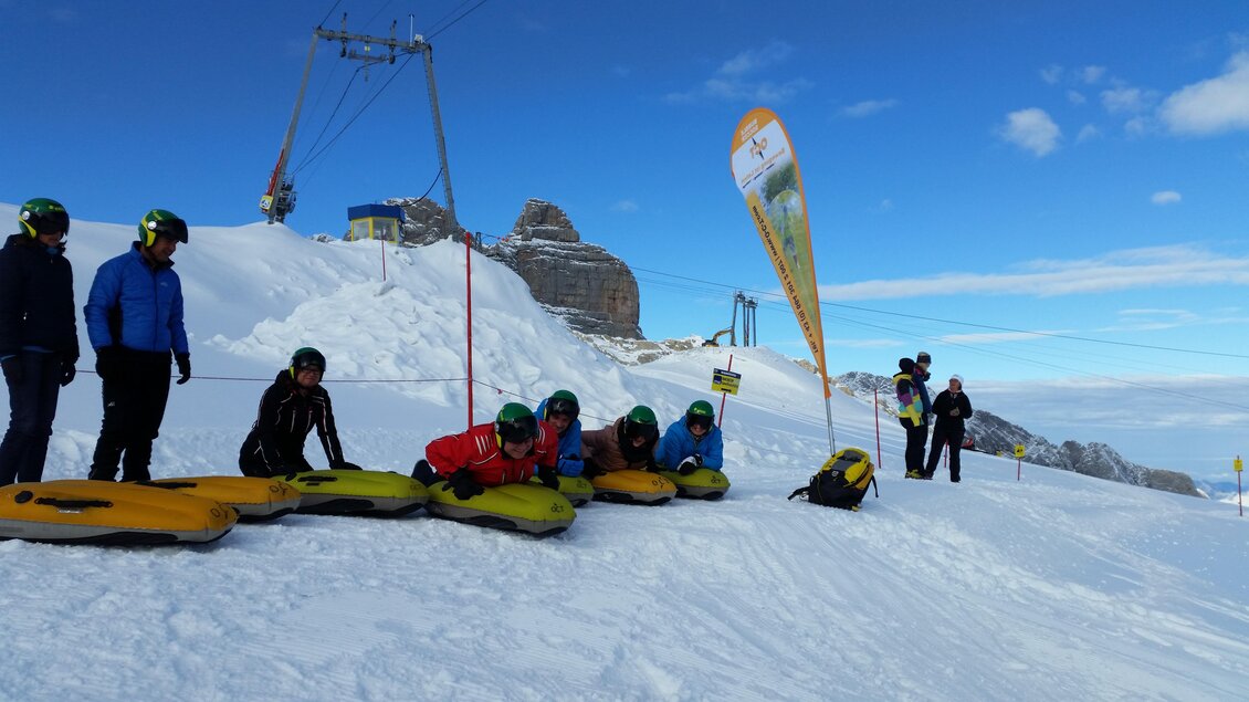 Eine Gruppe von Menschen in winterlicher Kleidung liegt auf Rutschschlitzen auf schneebedecktem Gelände. Im Hintergrund sind Berggipfel und ein blauer Himmel zu sehen. | © Thomas Humer