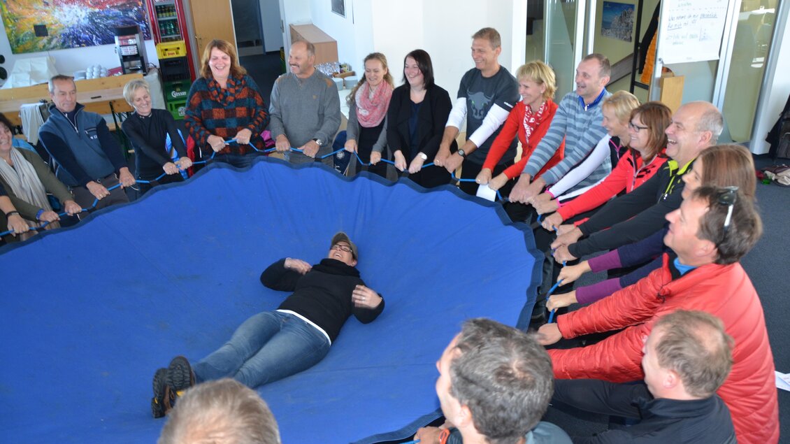 Eine Gruppe von Menschen sitzt im Kreis und hält einen großen blauen Stoff in der Mitte. Eine Person liegt auf dem Stoff und blickt entspannt auf die anderen. | © Thomas Humer