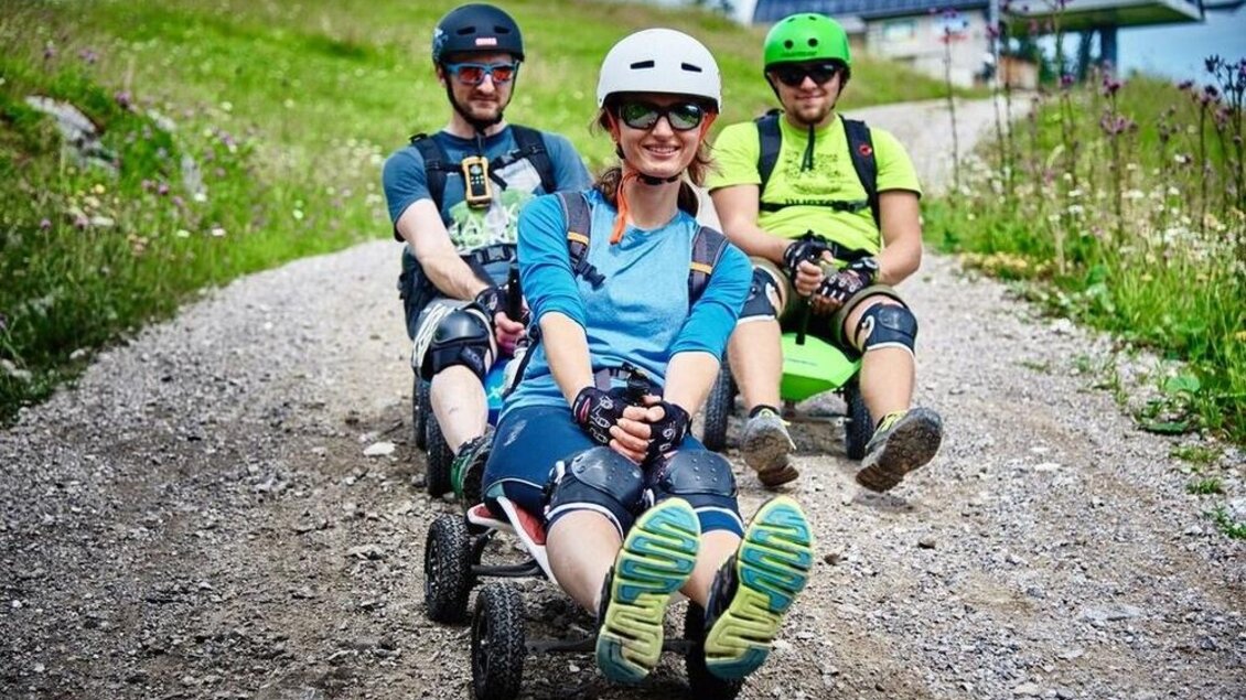 Drei Personen fahren auf einem praktischen Rodel auf einem Schotterweg. Alle tragen Helme und Schutzkleidung und genießen das Abenteuer in der Natur. | © Thomas Humer