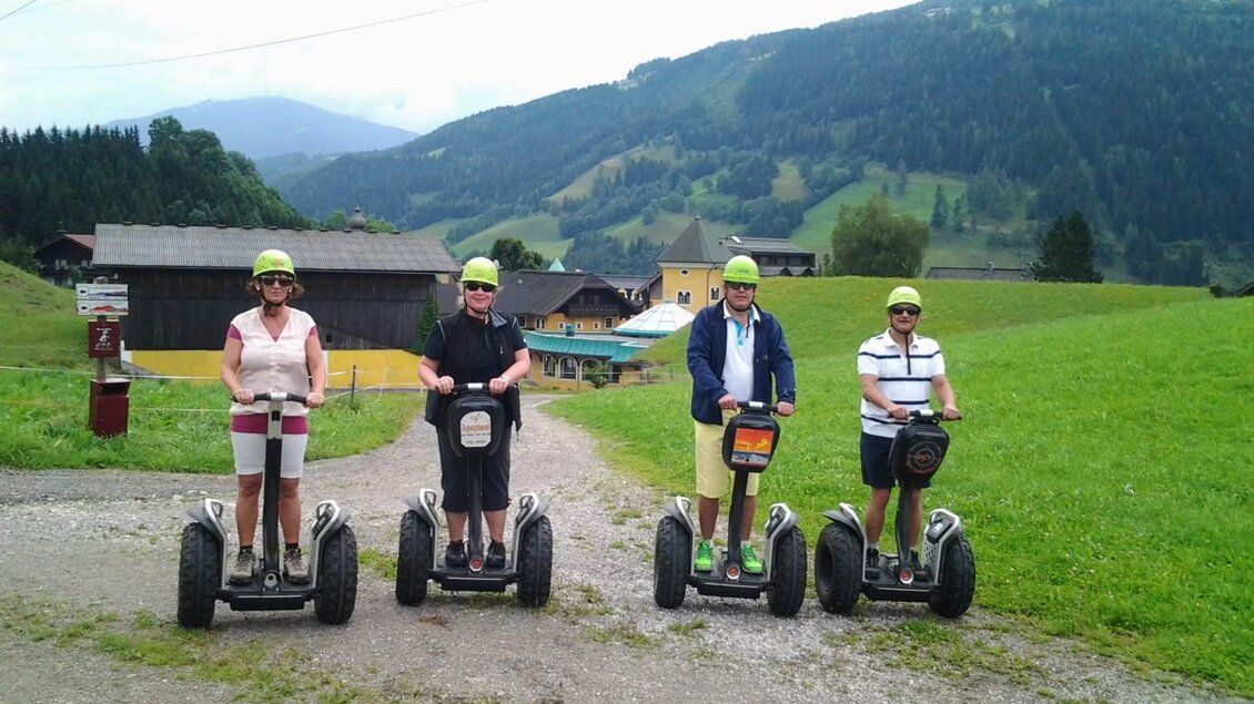 Vier Personen fahren auf Segways in einer grünen Landschaft mit Bergen im Hintergrund. Alle tragen Helme und stehen in einer Reihe auf einer Schotterstraße. | © Thomas Humer