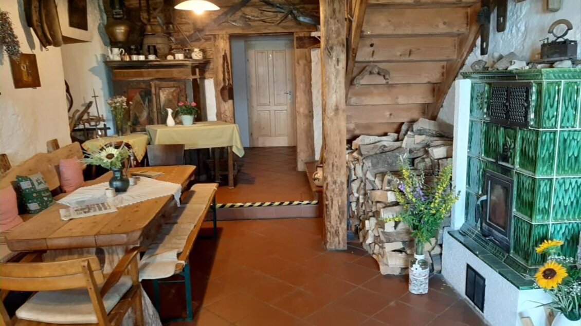 Ein gemütlicher Wohnraum mit Holztreppe und rustikalem Dekor. Im Raum befinden sich ein Tisch mit Stühlen, ein Ofen und ein Stapel Holz. | © Oberkogler-Alm