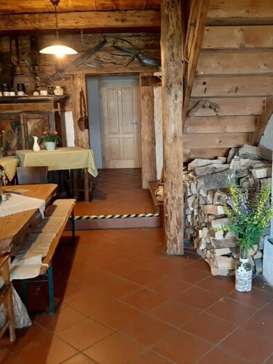 A cozy living space with a wooden staircase and rustic decor. The room contains a table with chairs, a stove, and a stack of wood. | © Oberkogler-Alm