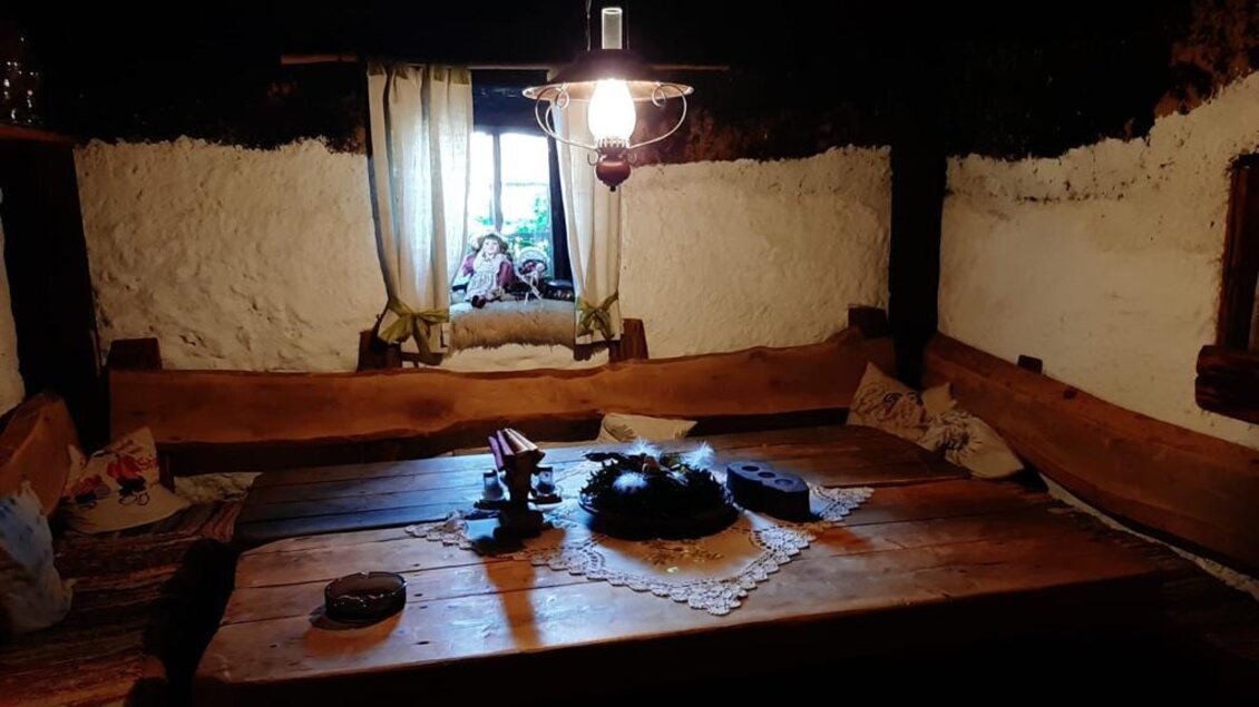 Ein rustikales Zimmer mit einem großen Holztisch in der Mitte. Das Licht einer Laterne beleuchtet den Raum, während ein Fenster den Blick nach draußen ermöglicht. | © Oberkogler-Alm