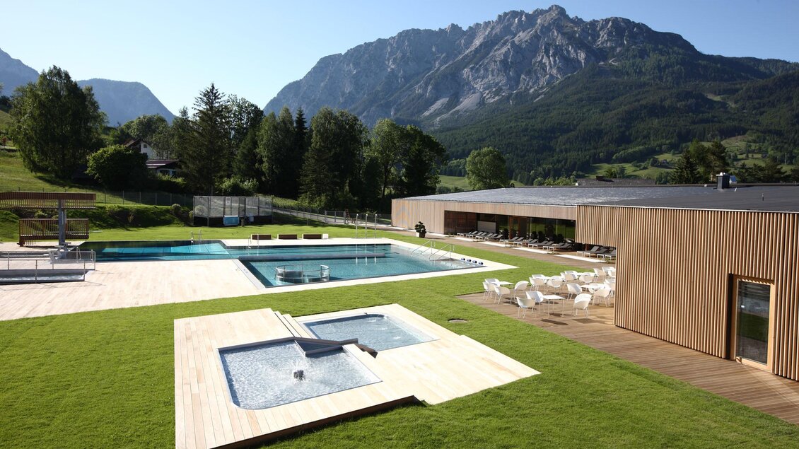 Ein modernes Schwimmbadgebiet mit mehreren Pools und einem Garten. Im Hintergrund erheben sich majestätische Berge unter einem klaren blauen Himmel. | © Panoramabad Gröbming 