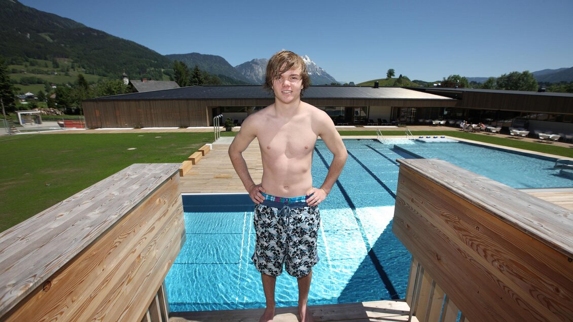 Ein junger Mann steht am Rand eines Schwimmbeckens auf einer Holzterrasse. Im Hintergrund sind Berge und ein modernes Gebäude sichtbar. | © Gemeinde Gröbming 