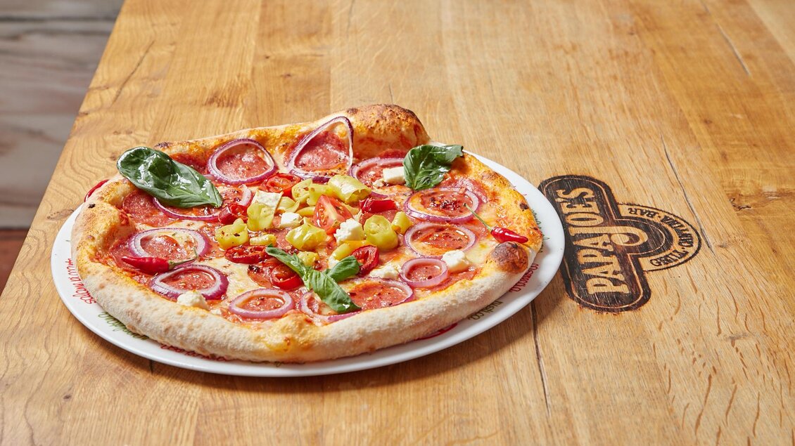 Eine frisch gebackene Pizza mit Tomaten, Zwiebeln und Basilikum auf einem Holzbrett. Das Bild zeigt eine appetitliche Präsentation der Pizza. | © PhotoINstyle - Brigit Steinberger