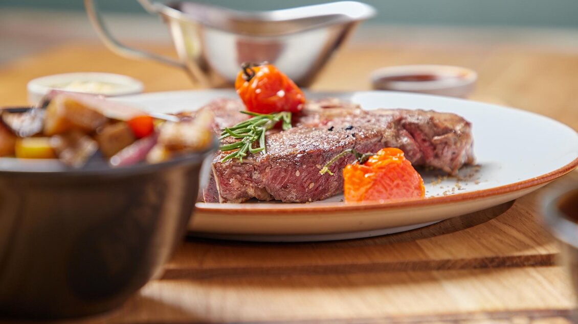 Ein zartes Steak mit gegrillten Tomaten und frischen Kräutern auf einem weißen Teller. Daneben befindet sich eine Schüssel mit buntem Gemüse. | © PhotoINstyle - Brigit Steinberger