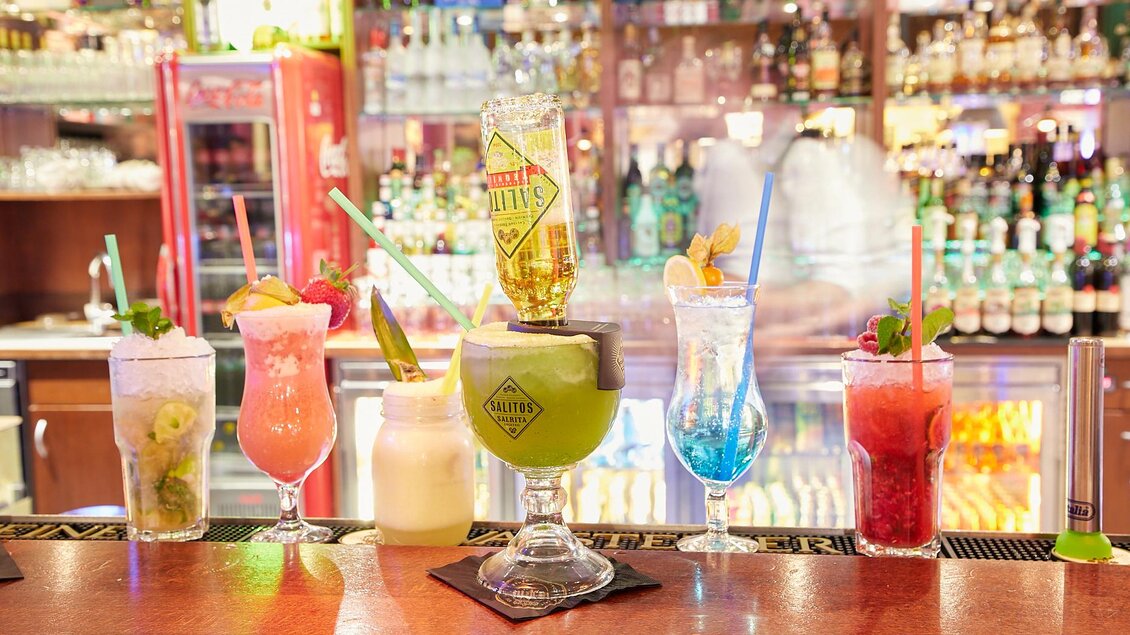 Bunte Cocktails stehen auf einer Bar, umgeben von einer Vielzahl von Spirituosen. Die Getränke sind kreativ dekoriert und laden zum Genießen ein. | © PhotoINstyle - Brigit Steinberger
