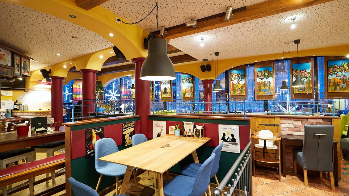 Eine gemütliche Restaurantumgebung mit bunten Fenstern und Holzdetails. Es gibt mehrere Tische und Stühle, die zum Verweilen einladen. | © PhotoINstyle - Brigit Steinberger