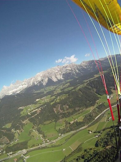A view from the air of a green landscape with mountains in the background. A paraglider with a yellow canopy and colorful lines is visible in the foreground. | © Flugschule Aufwind