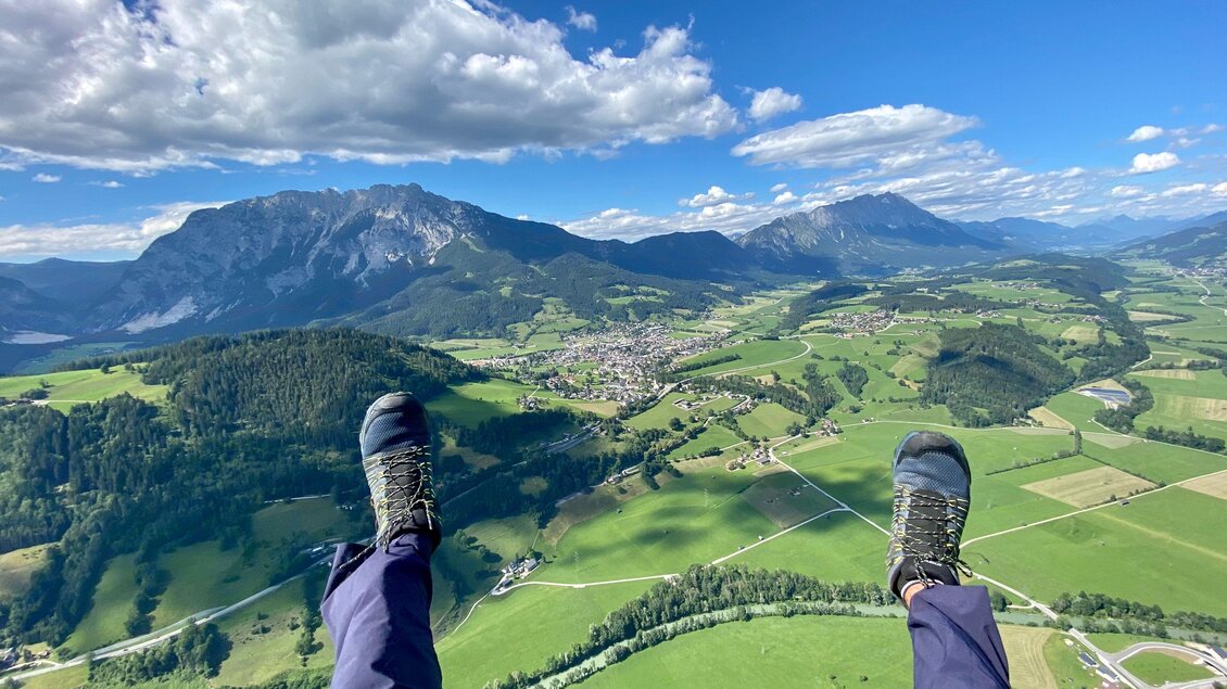 Ein Blick aus der Luft auf eine grüne Landschaft mit Bergen im Hintergrund. Zu sehen sind die Beine einer Person, die über der Landschaft schwebt. | © Sky Club Austria 