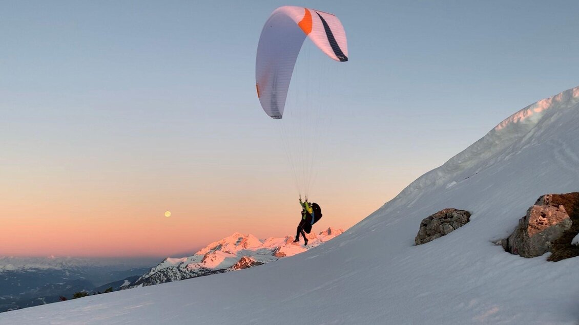 Ein Paraglider hebt in den Bergen ab, umgeben von schneebedeckten Gipfeln. Der Himmel hat einen sanften Farbverlauf bei Sonnenuntergang. | © Sky Club Austria 