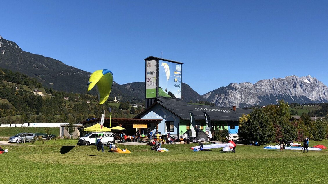 Ein malerischer Veranstaltungsort für Gleitschirmfliegen mit verschiedenen Gleitschirmen auf einer Wiese. Im Hintergrund erheben sich majestätische Berge unter einem klaren blauen Himmel. | © Sky Club Austria 