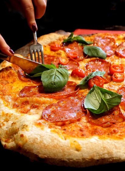 A freshly prepared pizza with salami, cherry tomatoes, and fresh basil. One person is cutting the pizza with a knife and fork. | © Symbolfoto