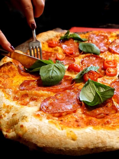 A freshly prepared pizza with salami, cherry tomatoes, and fresh basil. One person is cutting the pizza with a knife and fork. | © Symbolfoto