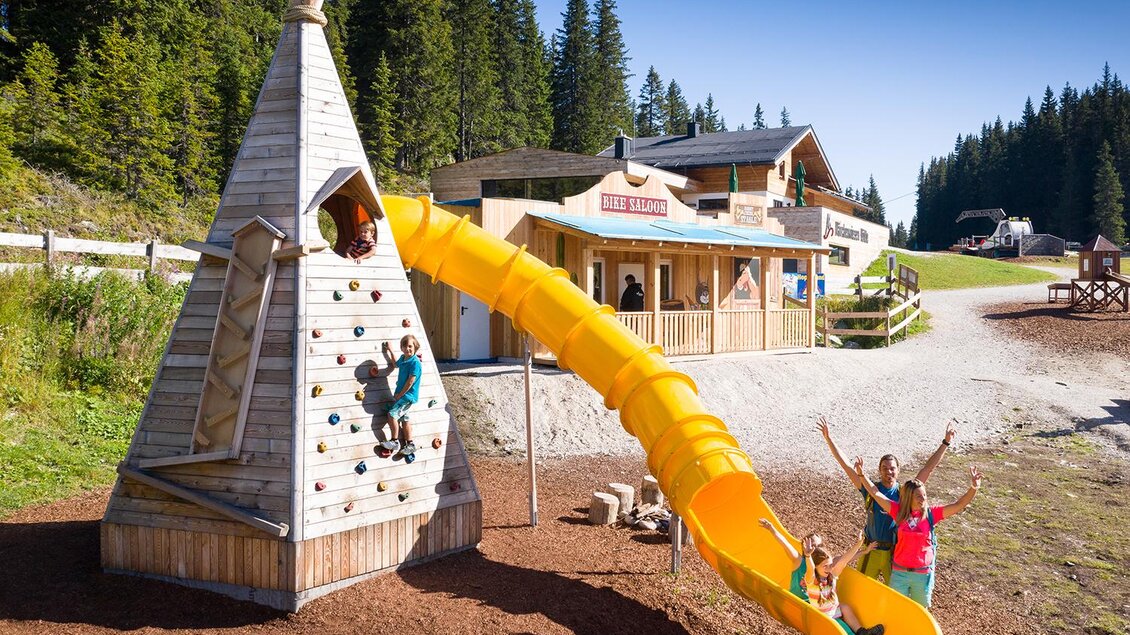 Ein Spielplatz mit einem Kletterturm in Form eines Tipis und einer gelben Rutsche. Kinder spielen fröhlich im Freien vor einer malerischen Berglandschaft. | © Josh Absenger