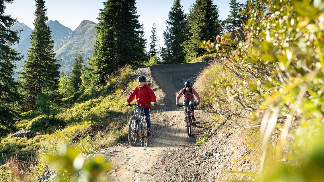 Zwei Radfahrer fahren auf einem Waldweg in den Bergen. Umgeben von grünen Bäumen und einem klaren Himmel genießen sie die Natur. | © Josh Absenger
