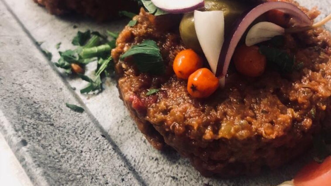 Zwei köstliche Patties, reich garniert mit frischen Kräutern, Zwiebeln und Oliven, serviert auf einem grauen Teller. Daneben sind farbenfrohe Beilagen zu sehen.