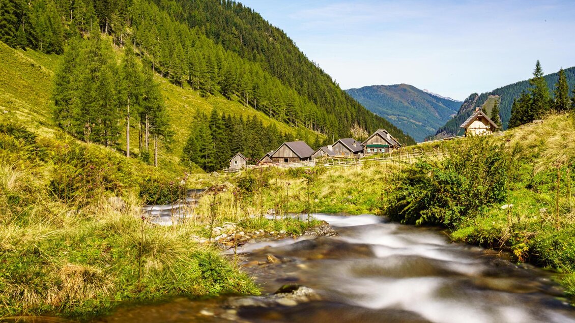 Ein malerisches Tal mit einer sanften Holzarchitektur und hohen grünen Bergen. Ein klarer Fluss fließt durch die Friedlichkeit der Landschaft. | © Posch Hof