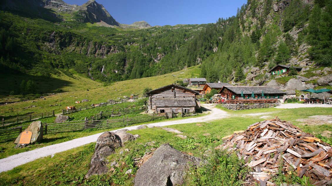 Eine malerische Alm in den Bergen mit Gebäude aus Holz und grünen Wiesen. Im Hintergrund erheben sich majestätische Berge und Bäume. | © Herbert Raffalt