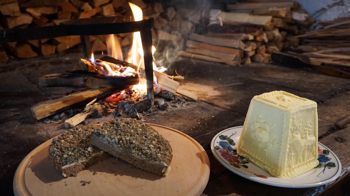 Ein rustikales Feuer in einem offenen Kamin, davor liegt ein Stück Brot auf einem Holzbrett. Neben dem Brot steht ein Stück Butter auf einem dekorativen Teller. | © Naturpark Sölktäler