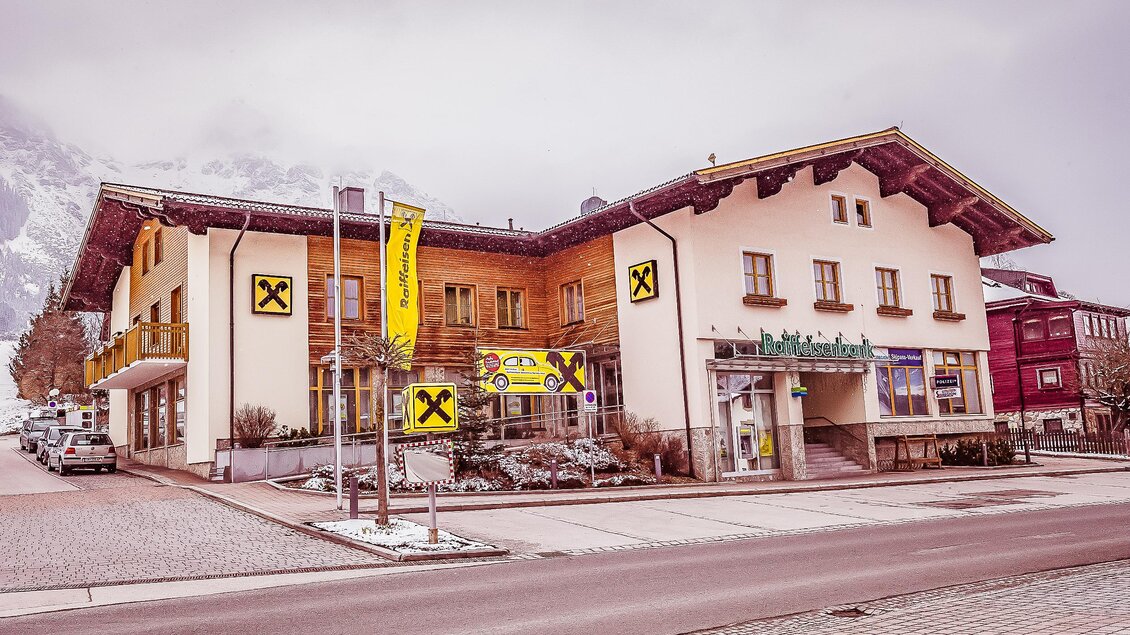 Ein modernes Gebäude mit Holzverkleidung und einem Geschäft im Erdgeschoss. Die Umgebung ist winterlich und ruhig, mit einigen Autos auf der Straße. | © Raiffeisenbank Schladming-Gröbming eGen