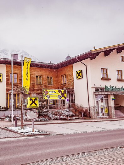A modern building with wooden cladding and a shop on the ground floor. The surroundings are wintry and quiet, with some cars on the street. | © Raiffeisenbank Schladming-Gröbming eGen
