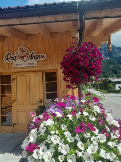 Ein Holzgebäude mit dem Schild "Red Angus" und blühenden Pflanzen in lila und weißen Farben. Im Hintergrund sind Berge und ein blauer Himmel zu sehen. | © Red Angus Ramsau