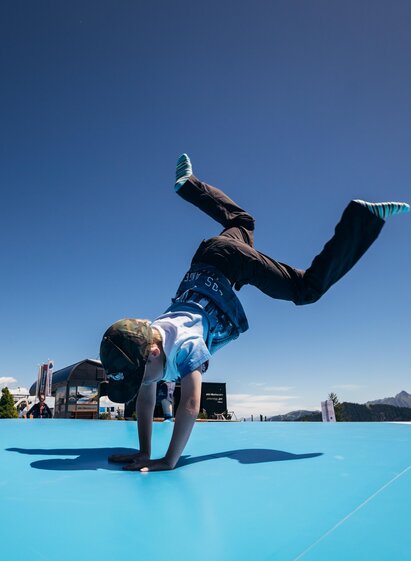 Ein Junge macht einen Handstand auf einer blauen Fläche. Im Hintergrund sind Berge und ein klarer Himmel zu sehen.