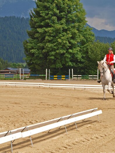 Eine Reiterin in roter Weste reitet auf einem weißen Pferd auf einem Sandplatz. Im Hintergrund sind Bäume und Berge zu sehen. | © Reiterhof Brandstätter