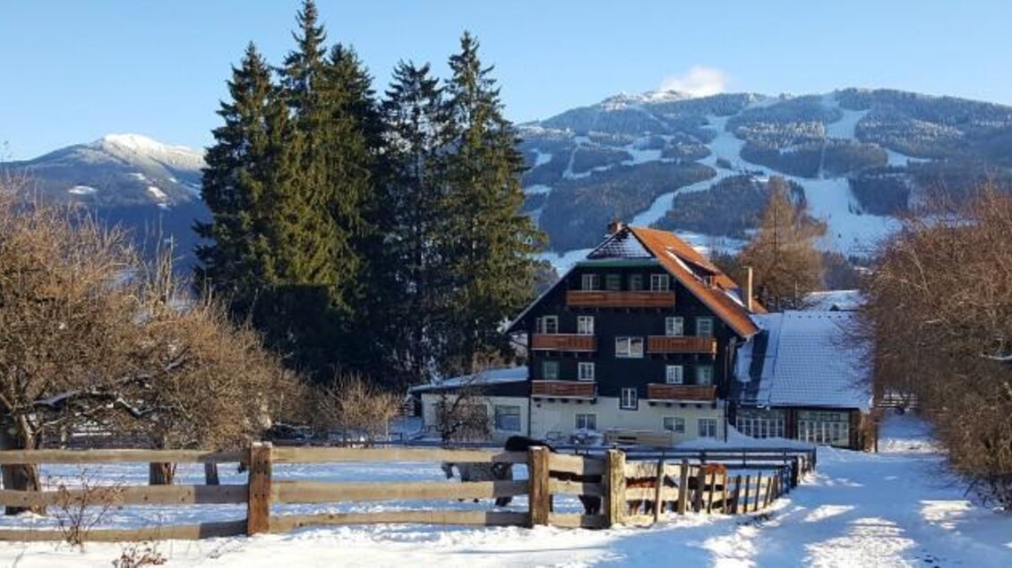 Ein gemütliches Haus im Schnee, umgeben von hohen Tannen und einer winterlichen Berglandschaft. Im Hintergrund sind schneebedeckte Pisten zu sehen. | © Reiterhof Brandstätter