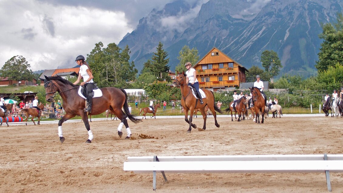 Reiter in einer Arena mit Bergen im Hintergrund. Die Teilnehmer zeigen ihre Pferde vor Zuschauern und einem rustikalen Gebäude. | © Reiterhof Brandstätter