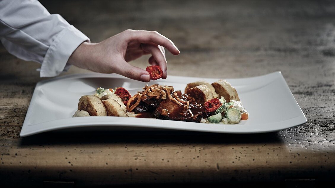 Ein eleganter Teller mit gourmet Gerichten, auf dem sich Fleisch, Beilagen und dekorative Elemente befinden. Eine Hand fügt gerade eine kleine Zutat hinzu. | © Hotel Pichlmayrgut