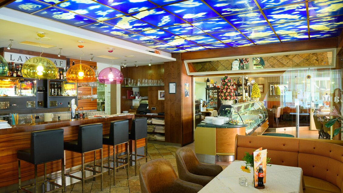 Ein modernes Restaurant mit einer stilvollen Bar und bunten Lampen. Im Hintergrund ist ein Eiswagen und eine gemütliche Sitzecke zu sehen. | © Restaurant-Cafe Landgraf