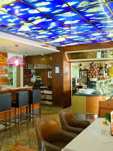 A modern restaurant with a stylish bar and colorful lamps. In the background, there is an ice cream cart and a cozy seating area. | © Restaurant-Cafe Landgraf