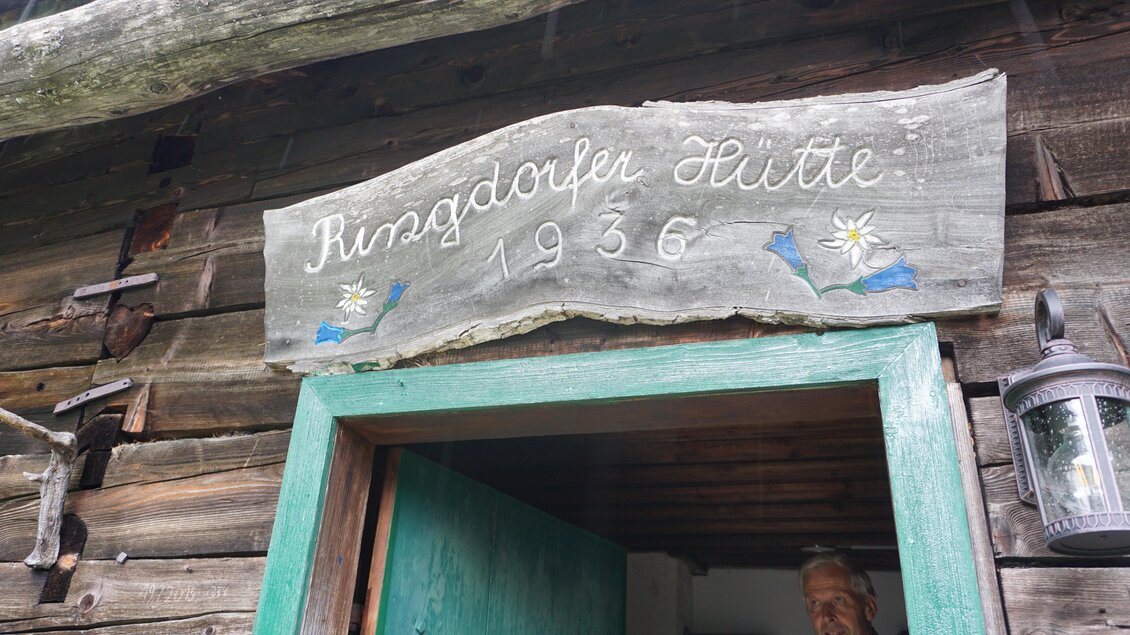 Ein Holzschild mit der Aufschrift "Ringdorfer Hütte 1936" über der Tür. Im Hintergrund ist eine Person zu sehen, die in die Kamera lächelt. | © Naturpark Sölktäler