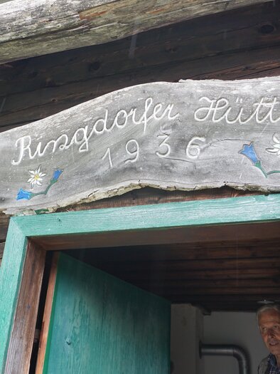 Ein Holzschild mit der Aufschrift "Ringdorfer Hütte 1936" über der Tür. Im Hintergrund ist eine Person zu sehen, die in die Kamera lächelt. | © Naturpark Sölktäler